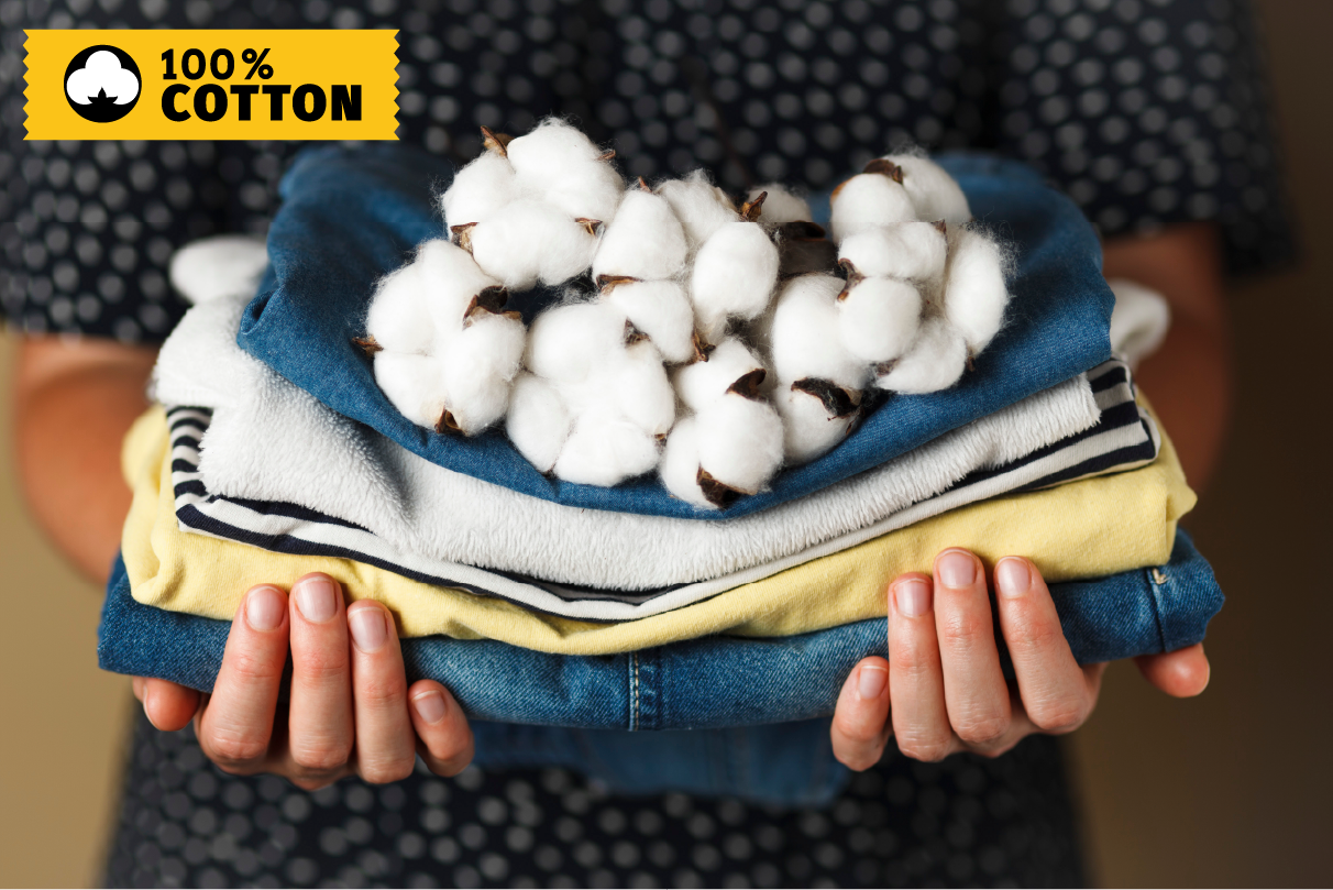 Stack of folded clothes held by a person with cotton displayed on top, labeled '100% Cotton'.