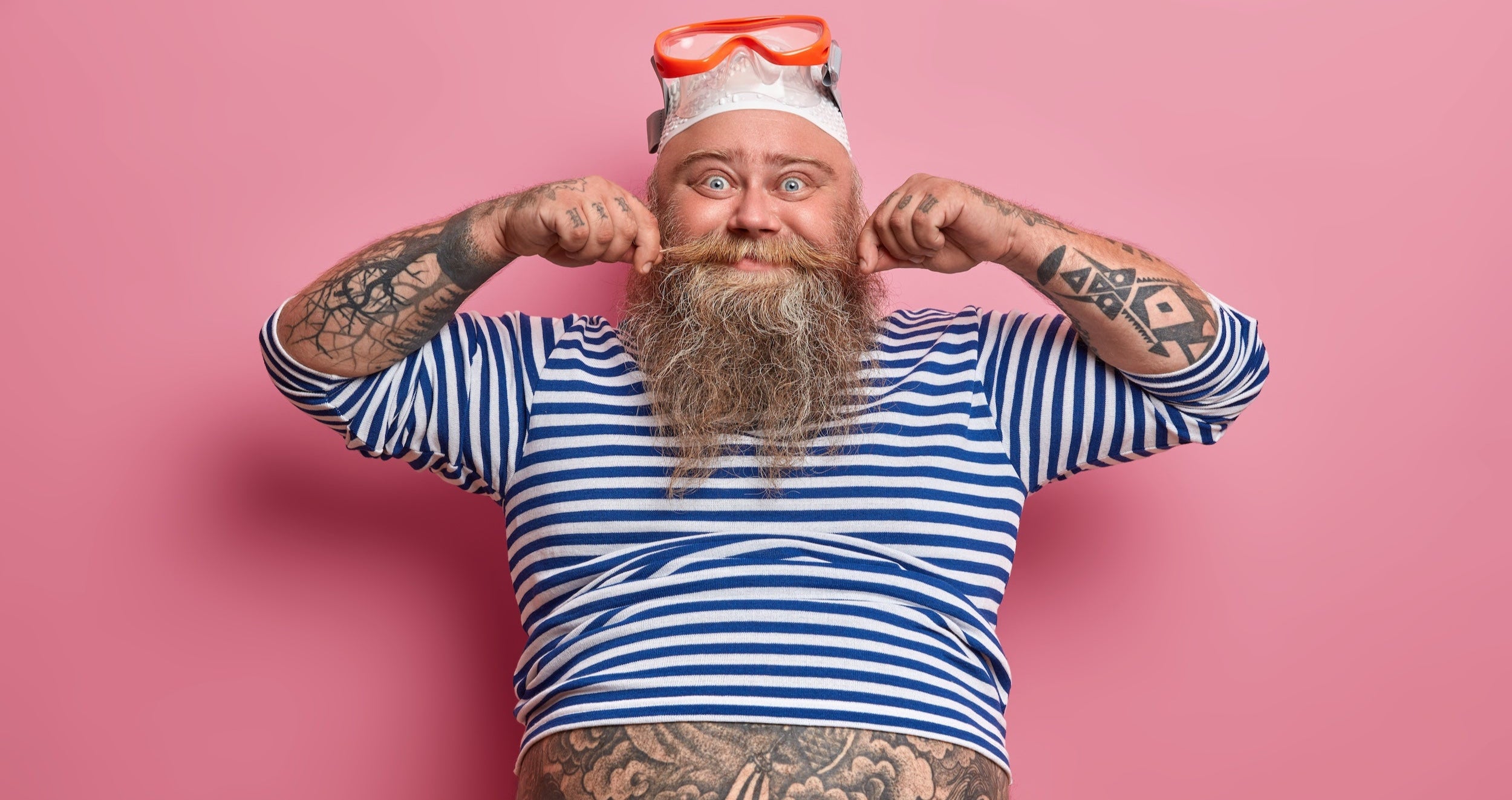 Man with tattoos and a beard wearing a striped shirt and diving goggles against a pink background