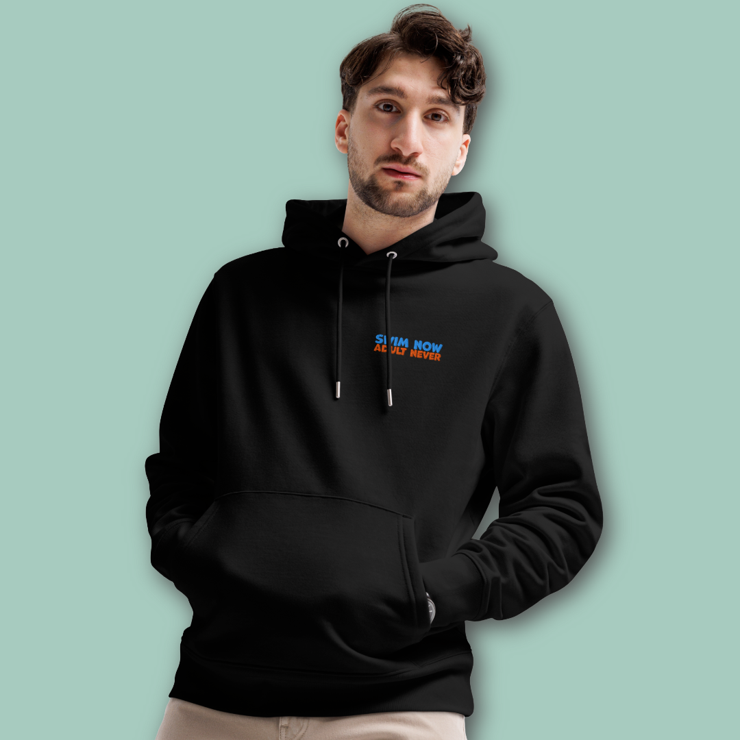 A man wearing a black organic hoodie by Lane Line Threads with a small blue and orange embroidered text "swim now adult never" on the left chest, standing against a solid light green background.