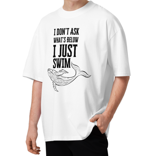 A young man with curly brown hair wearing a white organic oversized t-shirt with a black graphic print that reads 'I DON'T ASK WHAT'S BELOW I JUST SWIM' above a line drawing of a whale. He is standing against a solid white background.