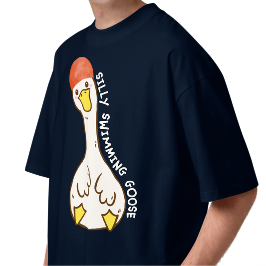 A young man with curly brown hair wearing a navy blue short-sleeve organic oversized t-shirt by Lane Line Threads featuring a cartoon graphic of a white swimming goose wearing a red swim cap, with the text 'SILLY SWIMMING GOOSE' printed vertically alongside it.