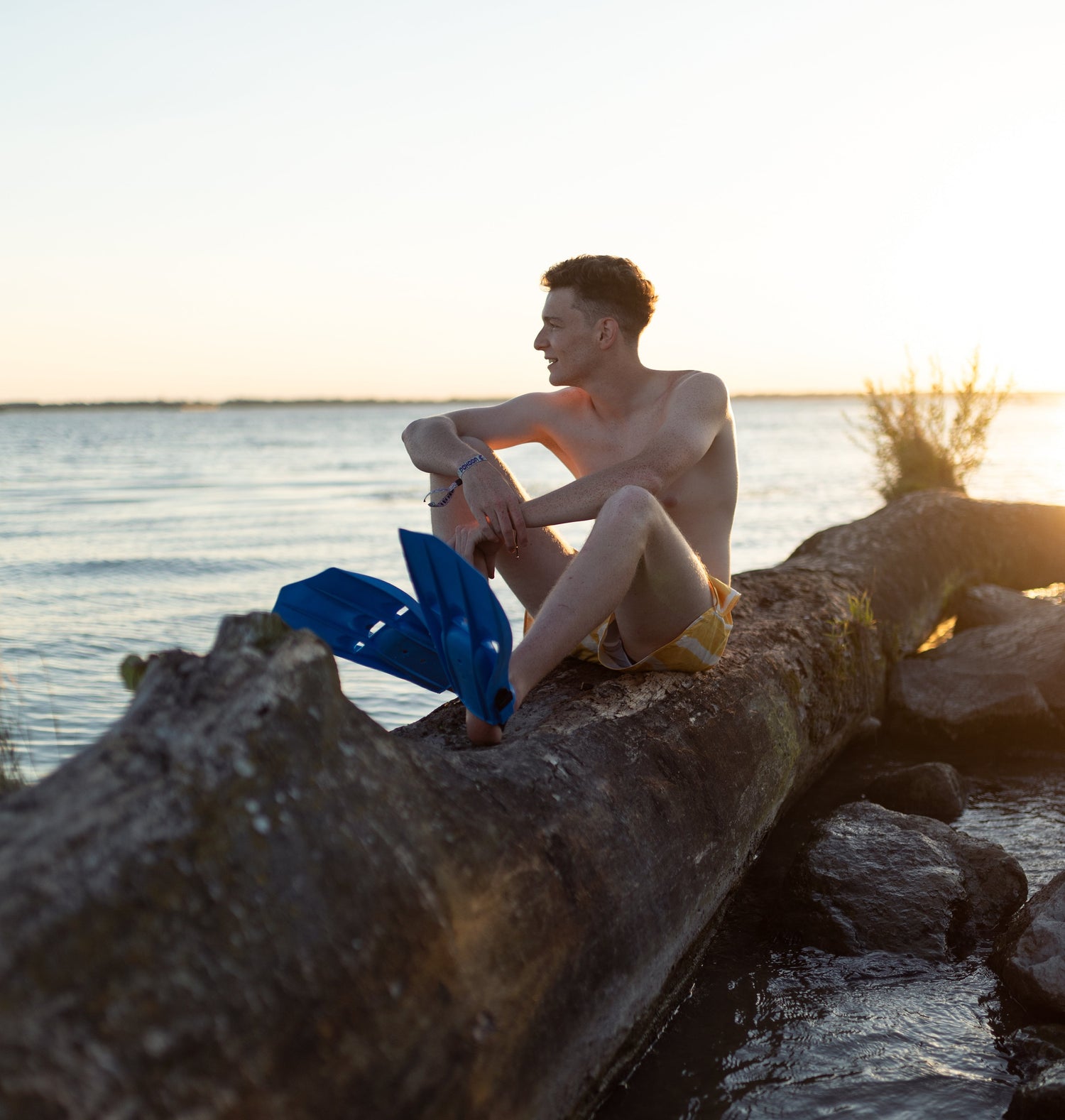 Man sitting on a log by the water with blue fins, during sunset.