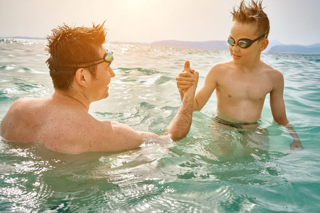 A man and a young boy, both wearing swim goggles, clasp hands in a firm "arm-wrestling" style handshake while standing in waist-deep ocean water.