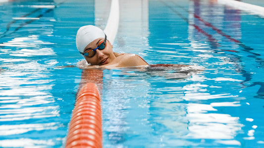 A swimmer wearing a white swim cap and goggles swims in a pool lane, with a bright orange divider rope running diagonally through the blue water talking about chlorine damaged hair