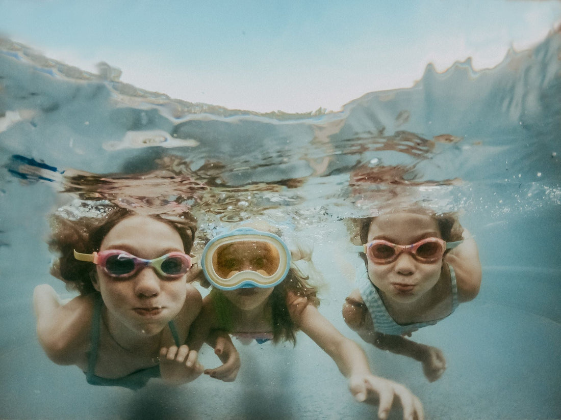 Three children swimming underwater with goggles, surrounded by clear blue water and sunlight.