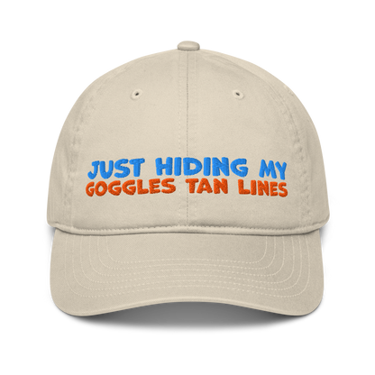Beige organic cotton baseball cap for swimmers with "JUST HIDING MY GOGGLES TAN LINES" embroidered in blue and orange text.
