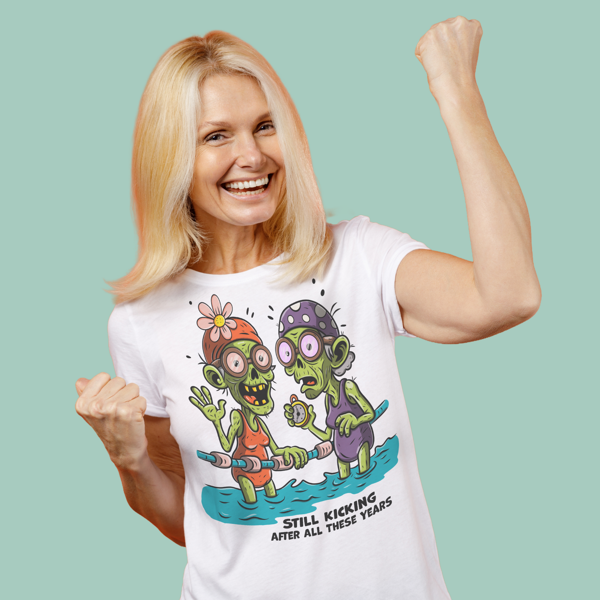 Person wearing a white organic Halloween swim shirt with a colorful cartoon illustration of two zombie-like swimmers standing in water, each holding a pool noodle. One wears an orange swimsuit and flowered swim cap; the other wears a purple swimsuit and polka-dotted cap. Both have exaggerated expressions and bulging eyes. Below the graphic, bold text reads: “STILL KICKING AFTER ALL THESE YEARS.”