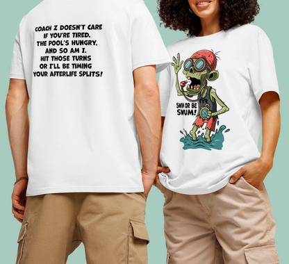 Two people wearing white organic swim-themed t-shirts. The person on the left faces away, showing bold black text on the back: “COACH Z DOESN’T CARE IF YOU’RE TIRED…” with a zombie-themed swim coach message. The person on the right faces forward, displaying a colorful cartoon zombie swim coach in swim gear with the phrase “SWIM OR BE SWUM!” below.