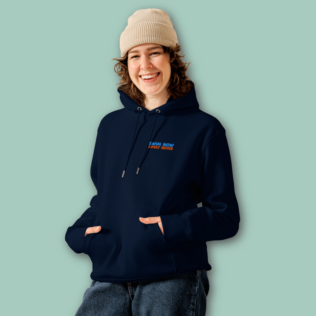 A woman wearing a navy-blue organic hoodie with a small blue and orange embroidered text "swim now adult never" on the left chest, paired with a beige knit beanie and dark jeans, standing against a solid light green background.