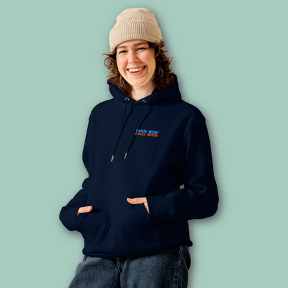 A woman wearing a navy-blue organic hoodie with a small blue and orange embroidered text "swim now adult never" on the left chest, paired with a beige knit beanie and dark jeans, standing against a solid light green background.