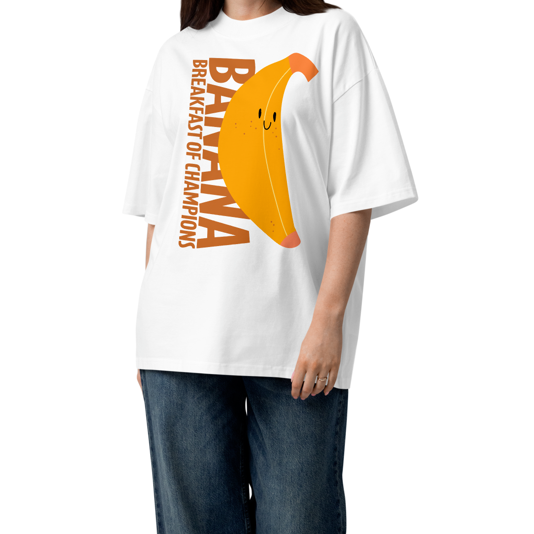 A woman modeling a white short-sleeve crewneck organic oversized t-shirt by Lane Line Threads featuring a graphic print of a smiling yellow banana with the text 'BANANA BREAKFAST OF CHAMPIONS' in brown. She is wearing wide-leg blue jeans and brown sandals against a plain white background.