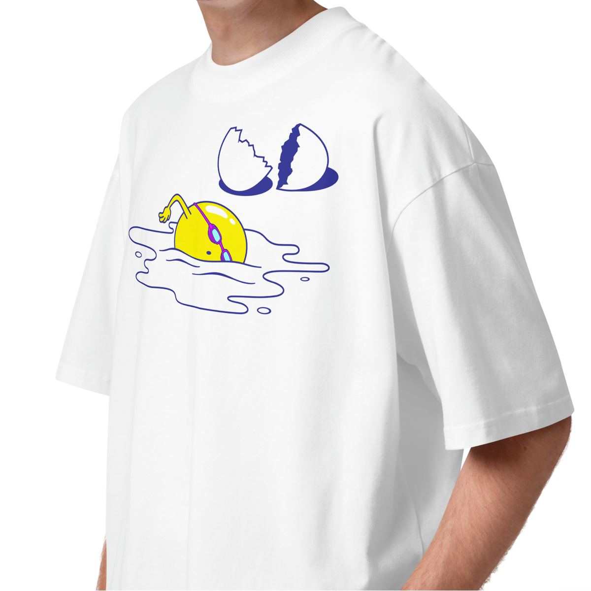 A young man with curly brown hair wearing a white short-sleeve crewneck organic oversized t-shirt by Lane Line Threads featuring a graphic print of a yellow cartoon egg yolk swimmer character breaking an egg on a white background.