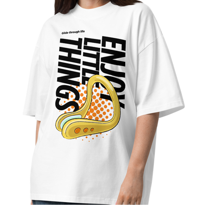 A young woman modeling a white short-sleeve crewneck organic oversized t-shirt by Lane Line Threads featuring a bold graphic print on the front. The design includes the text 'Enjoy Little Things' in black, a gold-colored swimming nose clip illustration, and a halftone pattern in orange.