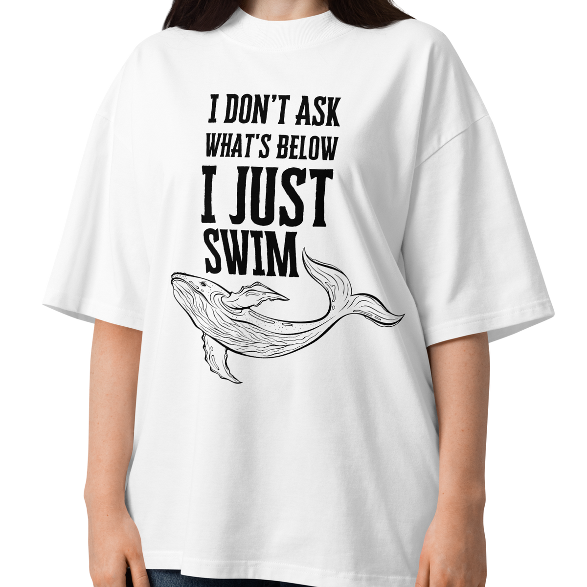 A woman modeling a white organic oversized t-shirt with a black graphic print that reads 'I DON'T ASK WHAT'S BELOW I JUST SWIM' above a line art illustration of a whale. She is wearing blue jeans against a plain white background.