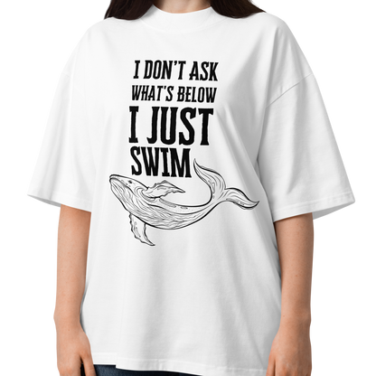 A woman modeling a white organic oversized t-shirt with a black graphic print that reads 'I DON'T ASK WHAT'S BELOW I JUST SWIM' above a line art illustration of a whale. She is wearing blue jeans against a plain white background.