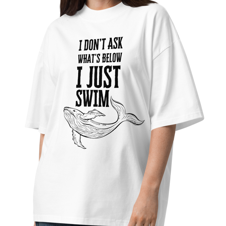 A woman modeling a white organic oversized t-shirt with the text 'I DON'T ASK WHAT'S BELOW I JUST SWIM' and a black line drawing of a whale, paired with dark blue wide-leg jeans and brown sandals, on a white studio background.