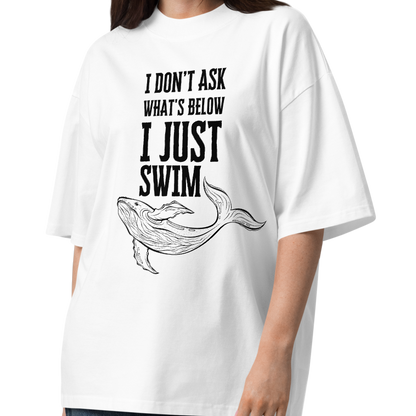 A woman modeling a white organic oversized t-shirt with the text 'I DON'T ASK WHAT'S BELOW I JUST SWIM' and a black line drawing of a whale, paired with dark blue wide-leg jeans and brown sandals, on a white studio background.