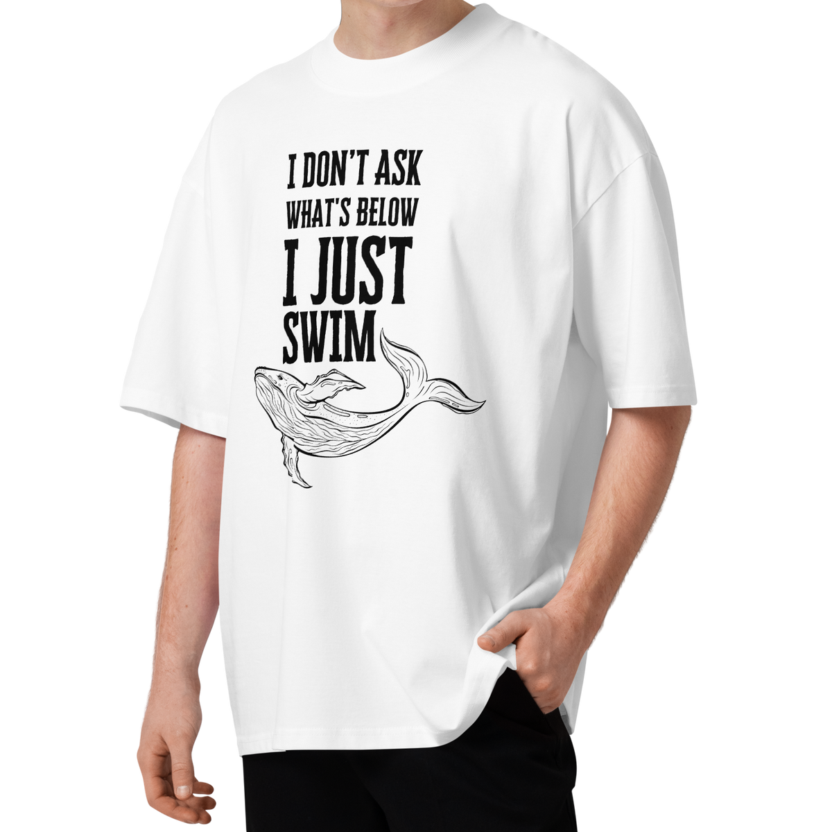 A young man with curly brown hair wearing a white organic oversized t-shirt with a black graphic print that reads 'I DON'T ASK WHAT'S BELOW I JUST SWIM' above a line drawing of a whale. He is standing against a solid white background.