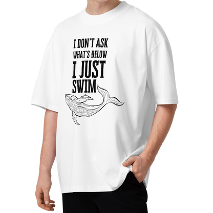 A young man with curly brown hair wearing a white organic oversized t-shirt with a black graphic print that reads 'I DON'T ASK WHAT'S BELOW I JUST SWIM' above a line drawing of a whale. He is standing against a solid white background.