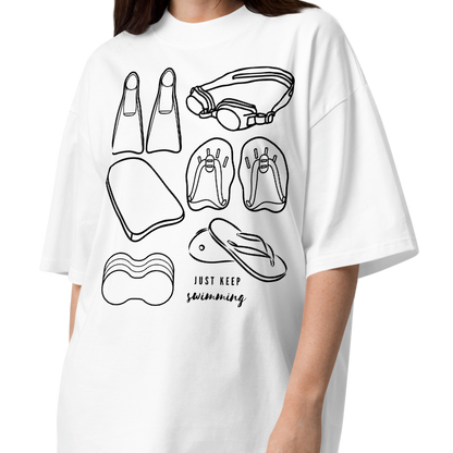 Person wearing a short-sleeve organic oversized t-shirt by Lane Line Threads with a black line art graphic of swimming gear, including flippers, goggles, paddles, kickboard and a flip-flop, with the text 'JUST KEEP swimming' printed on the front.