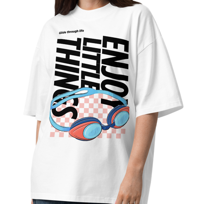 A woman modeling an organic oversized t-shirt in white with colorful swim goggles, bold “Enjoy Little Things” lettering, and “Glide Through Life” graphic by Lane Line Threads.