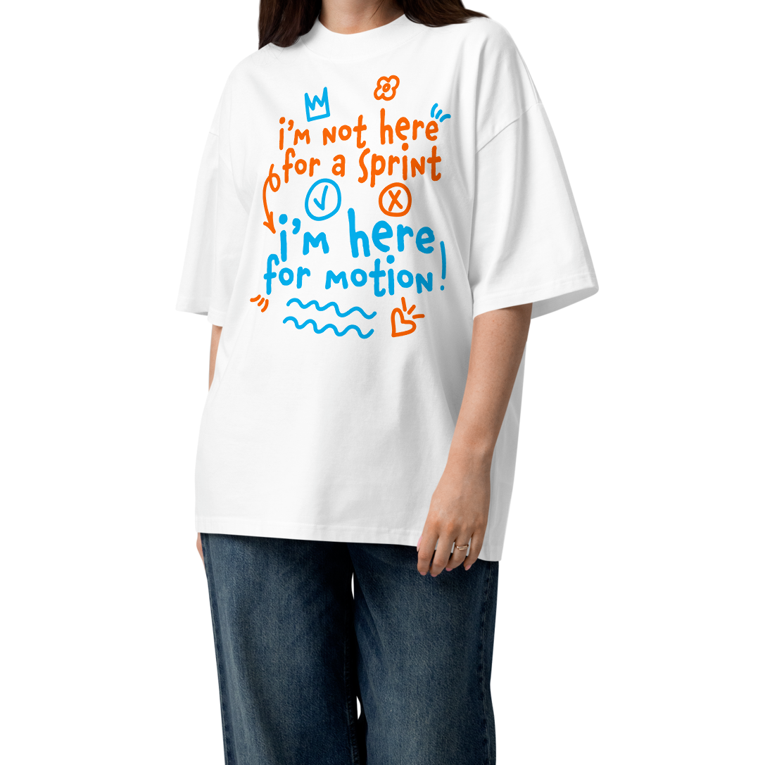 A woman modeling a white short-sleeve crewneck organic oversized t-shirt by Lane Line Threads with a colorful graphic print on the chest. The graphic features the text 'I'm not here for a sprint I'm here for motion!' in orange and blue handwritten-style lettering, accompanied by small doodles of a clock, a stop sign, wavy lines, and a heart. 