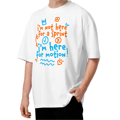 A young man with curly brown hair wearing a white short-sleeve crewneck organic oversized t-shirt by Lane Line Threads with a colorful graphic print on the chest. The graphic features the text 'I'm not here for a sprint I'm here for motion!' in orange and blue handwritten-style lettering, accompanied by small doodles of a clock, a stop sign, wavy lines, and a heart.