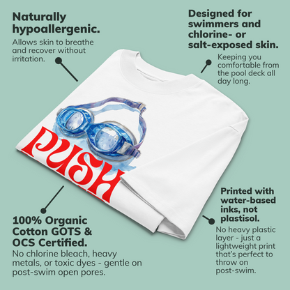 A folded white organic oversized t-shirt by Lane Line Threads with a large front graphic showing blue swim goggles and bold red 'PUSH' text, surrounded by marketing callouts on a pale green background. The text highlights hypoallergenic, swim-friendly, and certified cotton qualities.