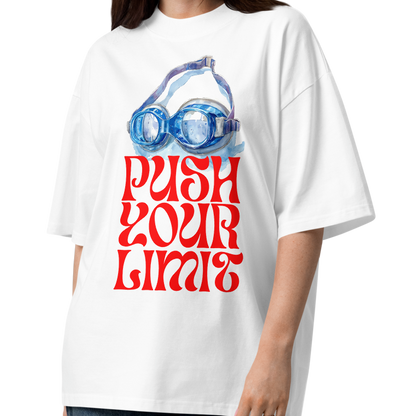 A woman modeling a white organic oversized t-shirt by Lane Line Threads with a retro graphic of swim goggles and the text 'PUSH YOUR LIMITS' in red, paired with wide-leg dark blue jeans and brown sandals, on a solid white background.