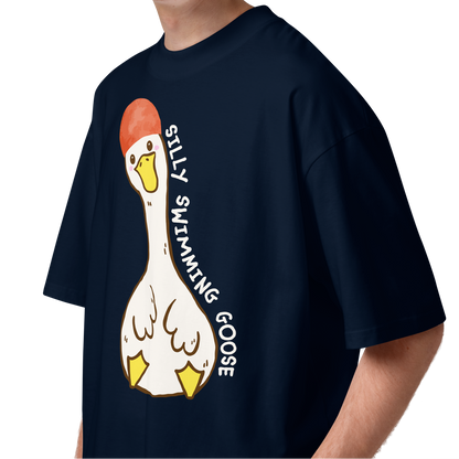 A young man with curly brown hair wearing a navy blue short-sleeve organic oversized t-shirt by Lane Line Threads featuring a cartoon graphic of a white swimming goose wearing a red swim cap, with the text 'SILLY SWIMMING GOOSE' printed vertically alongside it.