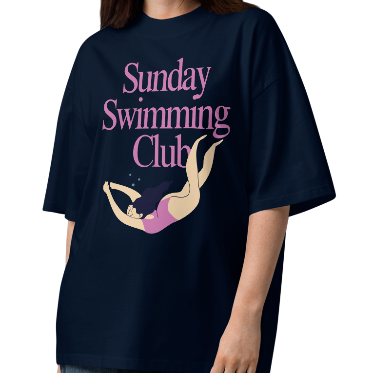 A woman modeling a navy-blue organic oversized t-shirt with a pink and yellow graphic that reads 'Sunday Swimming Club' on the chest, paired with loose-fit blue jeans and brown sandals, set against a plain white background.