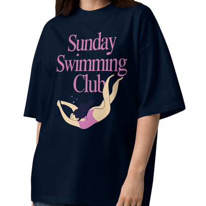 A woman modeling a navy-blue organic oversized t-shirt with a pink and yellow graphic that reads 'Sunday Swimming Club' on the chest, paired with loose-fit blue jeans and brown sandals, set against a plain white background.