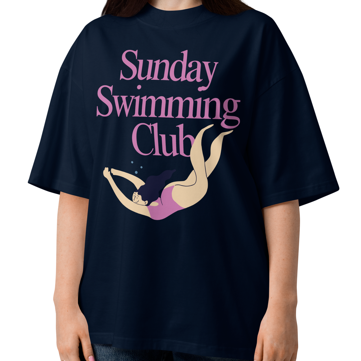 A woman modeling a navy-blue organic oversized t-shirt with a pink and beige graphic that reads 'Sunday Swimming Club' with an illustration of a girl swimmer.