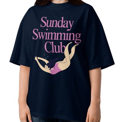 A woman modeling a navy-blue organic oversized t-shirt with a pink and beige graphic that reads 'Sunday Swimming Club' with an illustration of a girl swimmer.