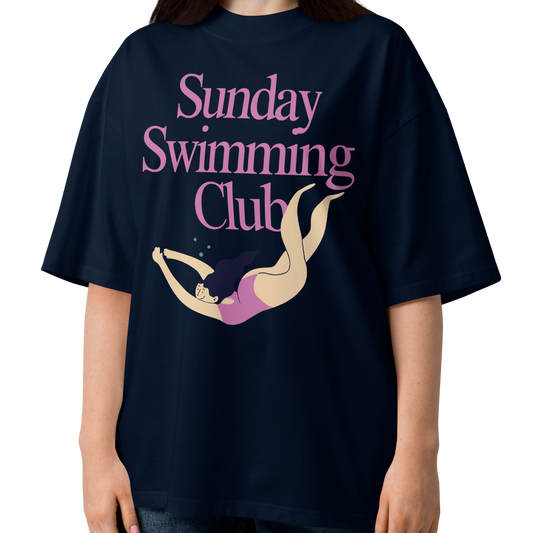 A woman modeling a navy-blue organic oversized t-shirt with a pink and beige graphic that reads 'Sunday Swimming Club' with an illustration of a girl swimmer.