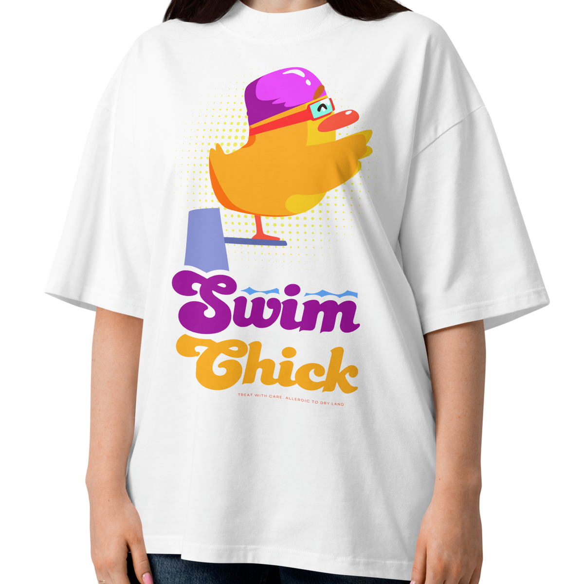A woman wearing a white short-sleeve crewneck organic oversized t-shirt by Lane Line Threads featuring a colorful graphic of a yellow duck wearing a purple swim cap and goggles, with the text 'Swim Chick' printed below in purple and orange, paired with dark blue jeans against a plain white background.