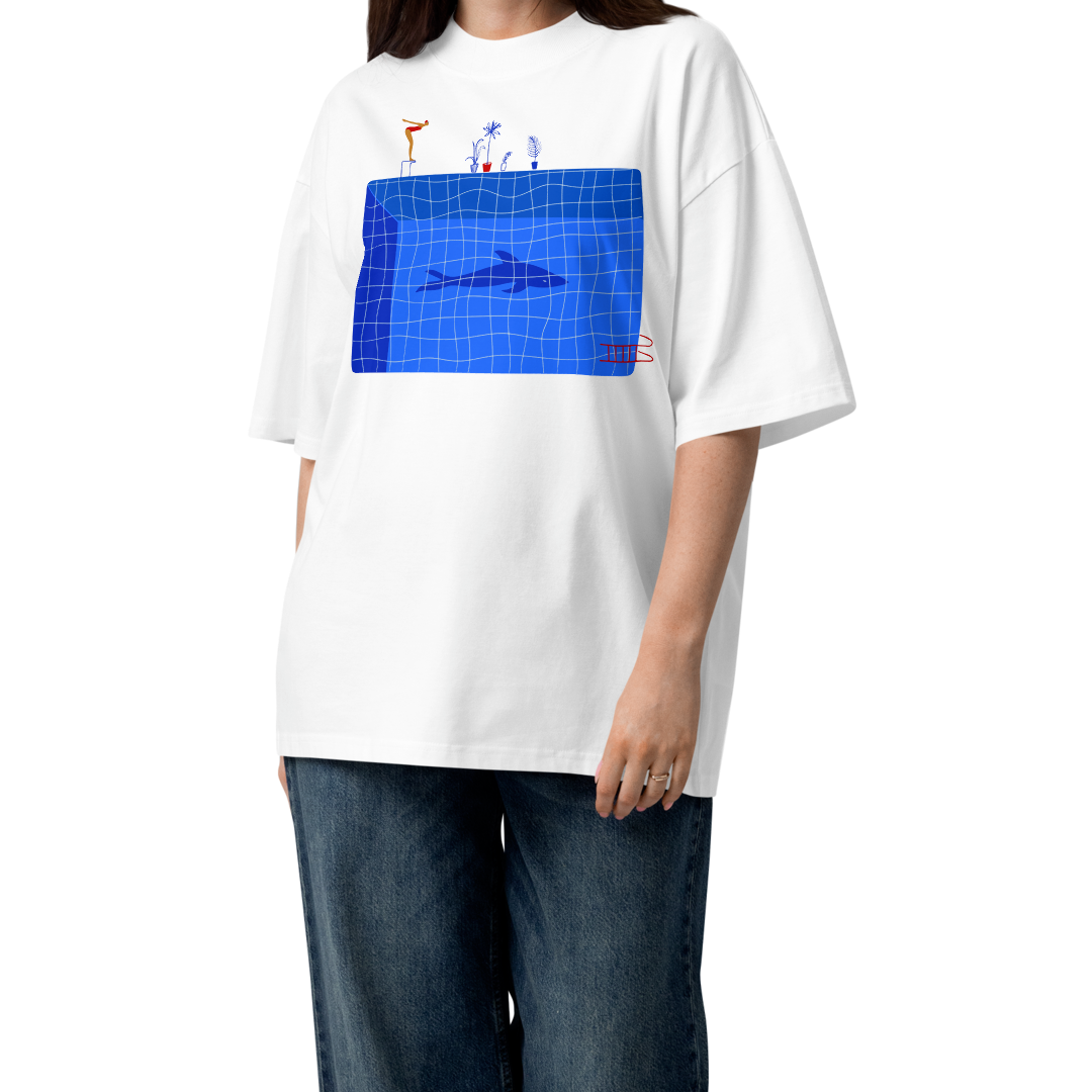 A young woman modeling a white short-sleeve crewneck organic oversized t-shirt by Lane Line Threads for swimmers featuring a graphic print of a blue swimming pool with a diving board, a diver, plants, and a shark on the front.