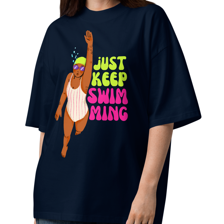A woman modeling a navy-blue organic oversized t-shirt with a cartoon girl swimmer and the text 'JUST KEEP SWIMMING' in green and pink, paired with wide-leg jeans and sandals on a white studio background.