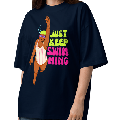 A woman modeling a navy-blue organic oversized t-shirt with a cartoon girl swimmer and the text 'JUST KEEP SWIMMING' in green and pink, paired with wide-leg jeans and sandals on a white studio background.