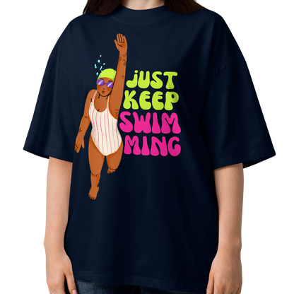 A woman modeling a navy-blue organic oversized t-shirt with a colorful graphic of a cartoon swimmer and the text 'JUST KEEP SWIM MING' in green, yellow, and pink, paired with blue jeans against a plain white background.