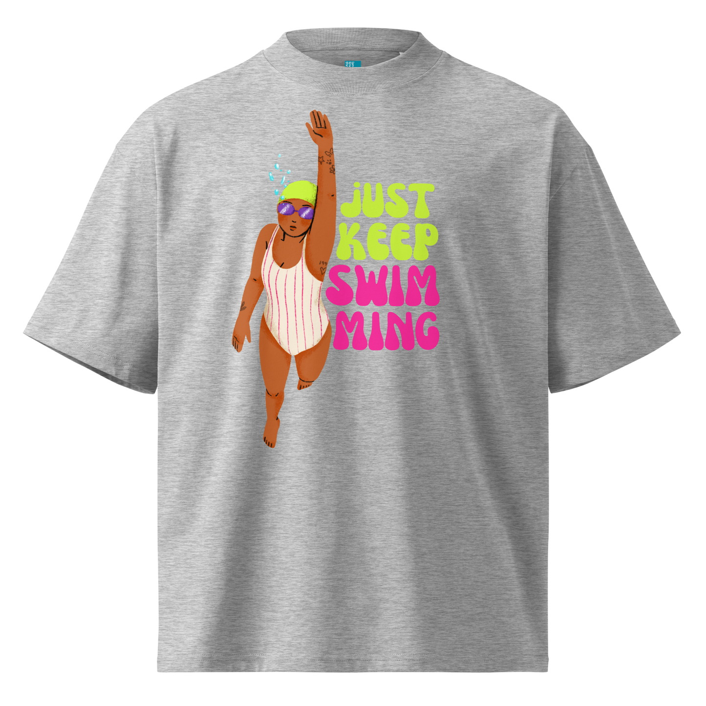 A heather gray short-sleeve organic oversized t-shirt featuring a graphic of a woman in a pink and white striped swimsuit with a green swim cap and purple goggles, next to the text 'JUST KEEP SWIMMING' in lime green and pink.