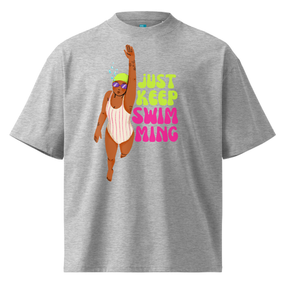 A heather gray short-sleeve organic oversized t-shirt featuring a graphic of a woman in a pink and white striped swimsuit with a green swim cap and purple goggles, next to the text 'JUST KEEP SWIMMING' in lime green and pink.