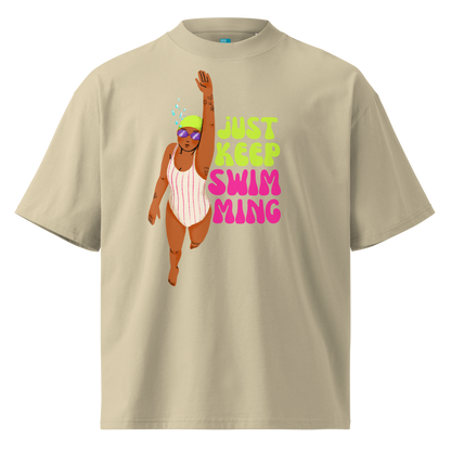 A beige short-sleeve crewneck organic oversized t-shirt featuring a graphic of a woman in a pink and white striped swimsuit with goggles, next to the text 'JUST KEEP SWIM MING' in neon green and pink.