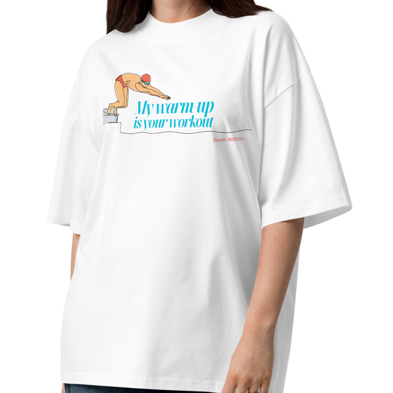 Woman wearing a white short-sleeve crewneck organic oversized t-shirt by Lane Line Threads featuring a graphic of a swimmer on a diving block with the text 'My warm-up is your workout' in blue and 'Swim Nation' in red below, displayed on a clean white background.
