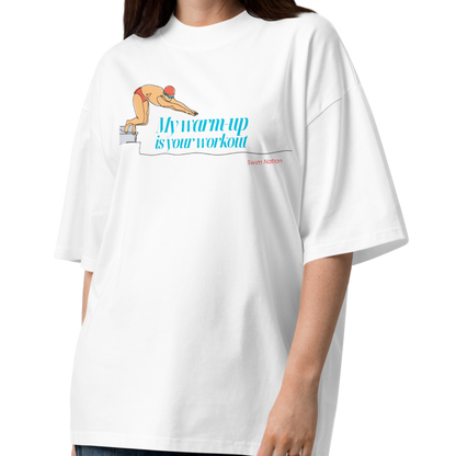 Woman wearing a white short-sleeve crewneck organic oversized t-shirt by Lane Line Threads featuring a graphic of a swimmer on a diving block with the text 'My warm-up is your workout' in blue and 'Swim Nation' in red below, displayed on a clean white background.