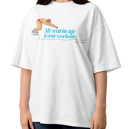A woman modeling a white short-sleeve crewneck organic oversized t-shirt by Lane Line Threads featuring a graphic of a swimmer on a diving block with the text 'My warm-up is your workout' in blue and 'Swim Nation' in red below, paired with dark blue jeans against a plain white background.