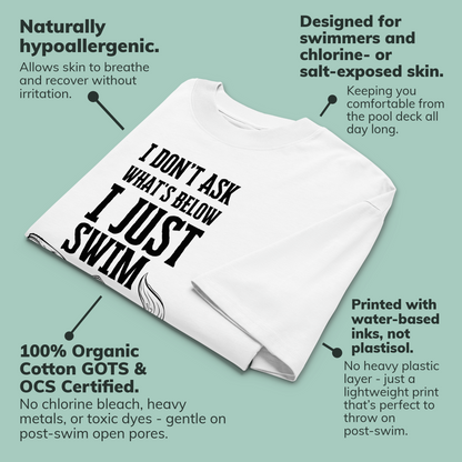 A white short-sleeve organic oversized t-shirt with large black text reading 'I DON'T ASK WHAT'S BELOW I JUST SWIM' on a light teal background with callout text describing product benefits.
