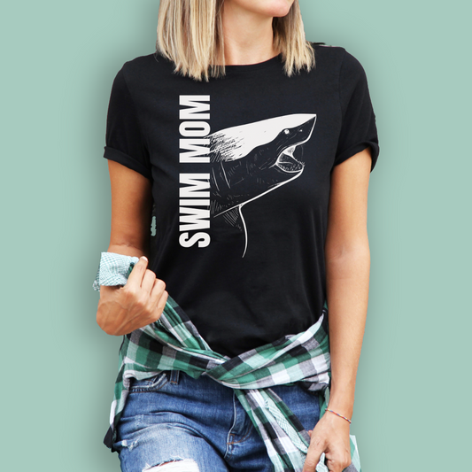 Person wearing a black organic swim mom shirt with a shark graphic and text against a light green background