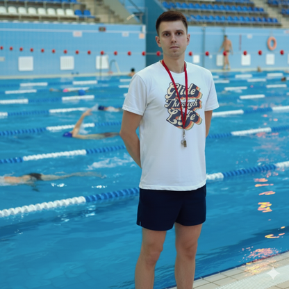 Swim coach wearing an organic swim shirt with text "glide through life" text on it, standing by a pool with swimmers in the background