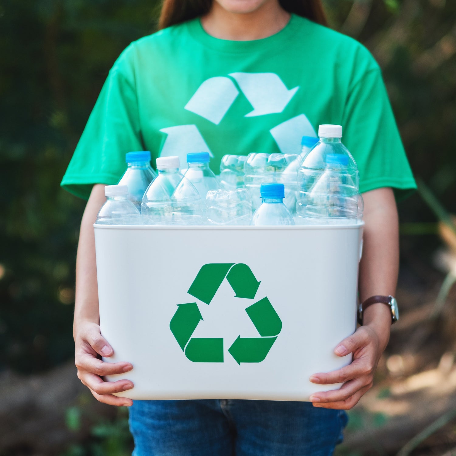 Person wearing a green shirt with a recycling symbol holding a box of recycled bottles.